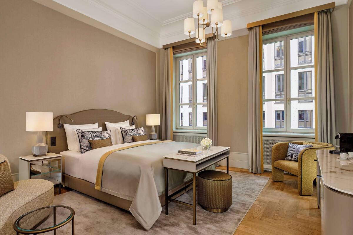 Ein Hotelzimmer mit einem großen Bett, zwei Nachttischen mit Lampen, einem kleinen runden Tisch, einem Sofa und Blick auf Gebäude durch große Fenster.