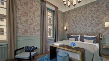 Quarto de hotel com cama, mesas de cabeceira, uma cadeira, janela com cortinas e um lustre.