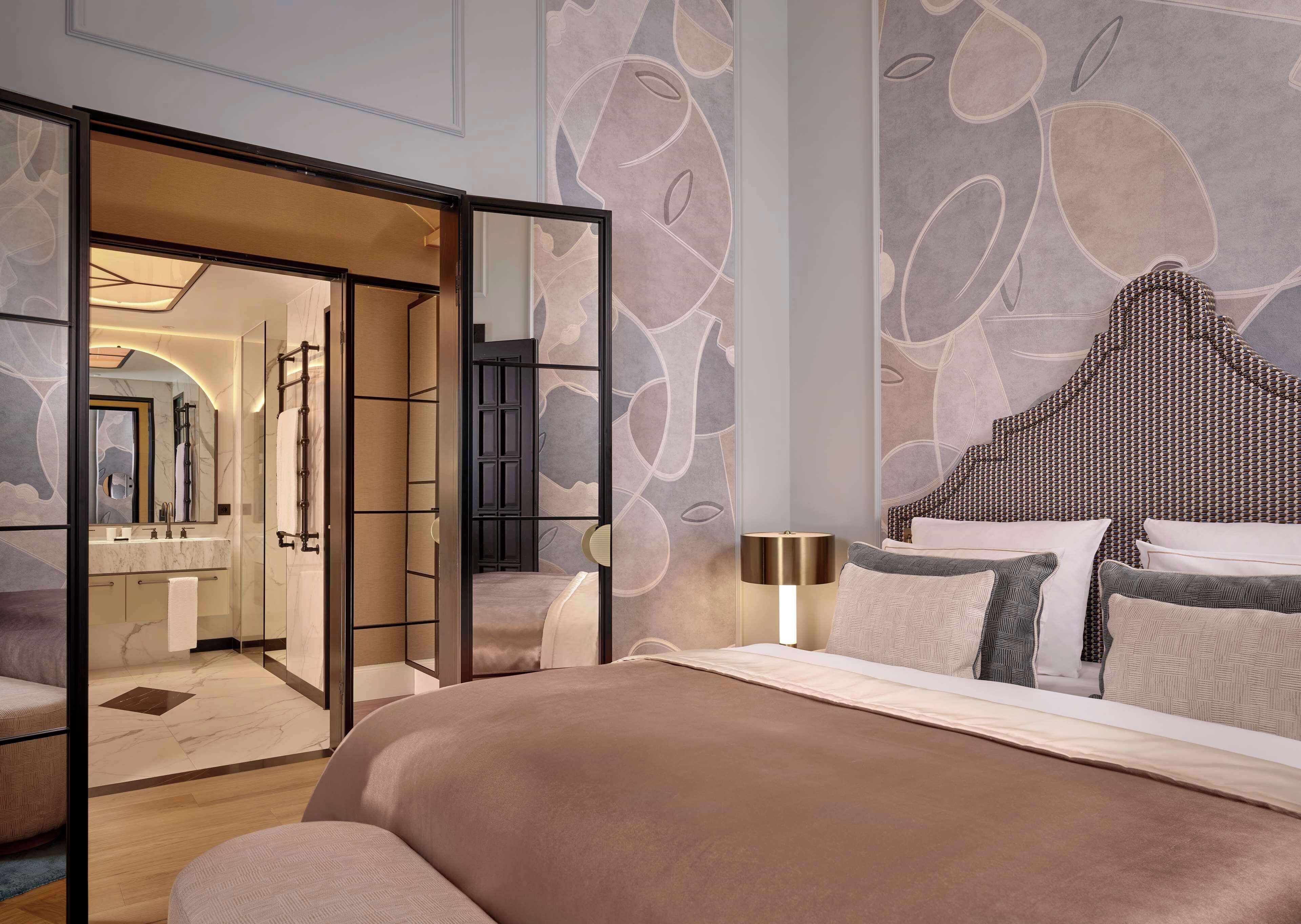 A modern hotel room with a large bed, a sitting area with a sofa, a bathroom with a vanity, and a large mirror.