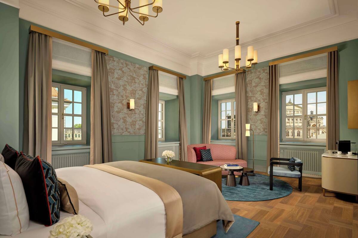 Ein Hotelzimmer mit einem großen Bett, einem Schreibtisch mit Stuhl, einem kleinen runden Tisch und Blick auf ein Gebäude durch die Fenster.