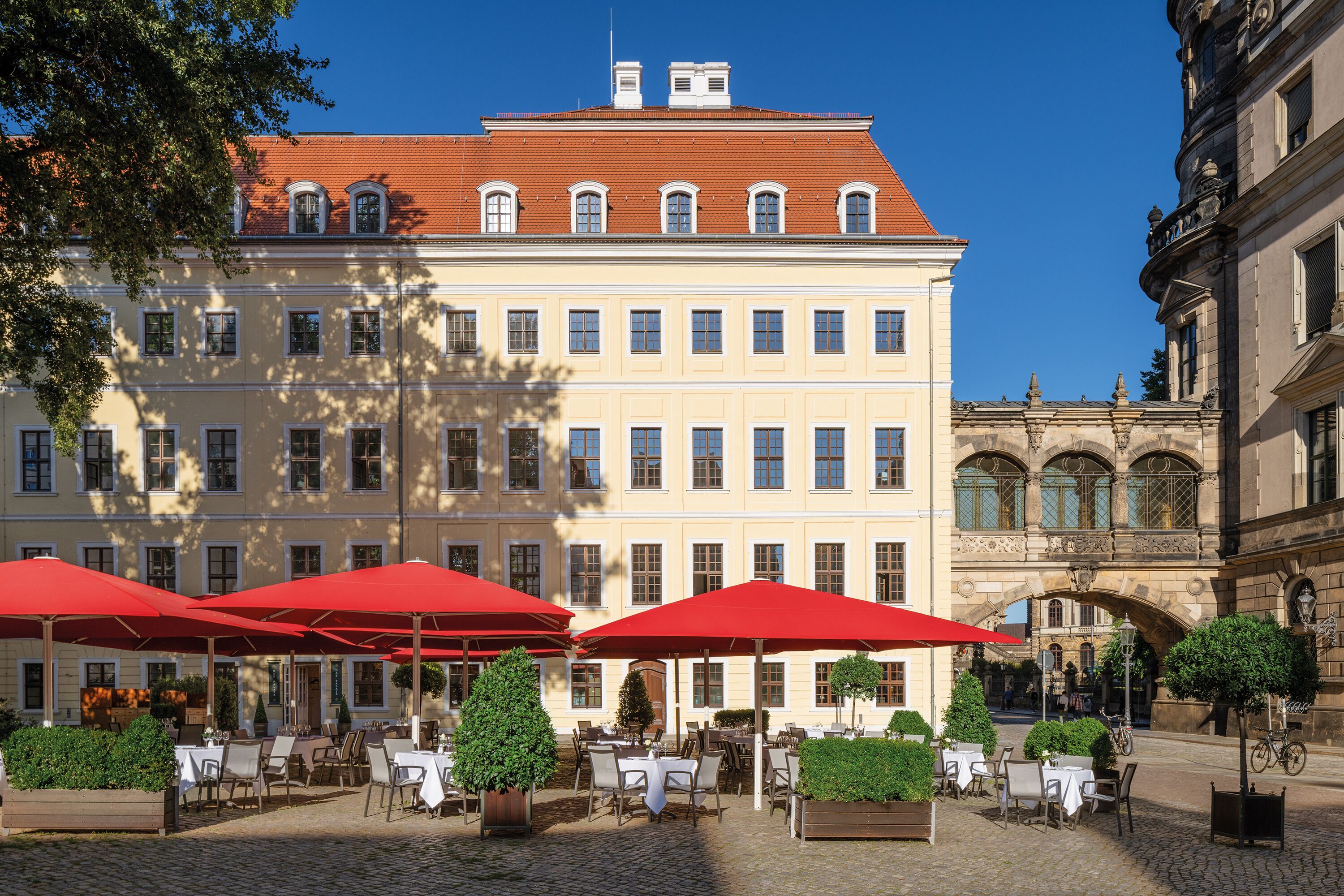 Ein Gebäude mit mehreren Fenstern und einem roten Dach. Draußen stehen Tische und Stühle mit roten Sonnenschirmen.