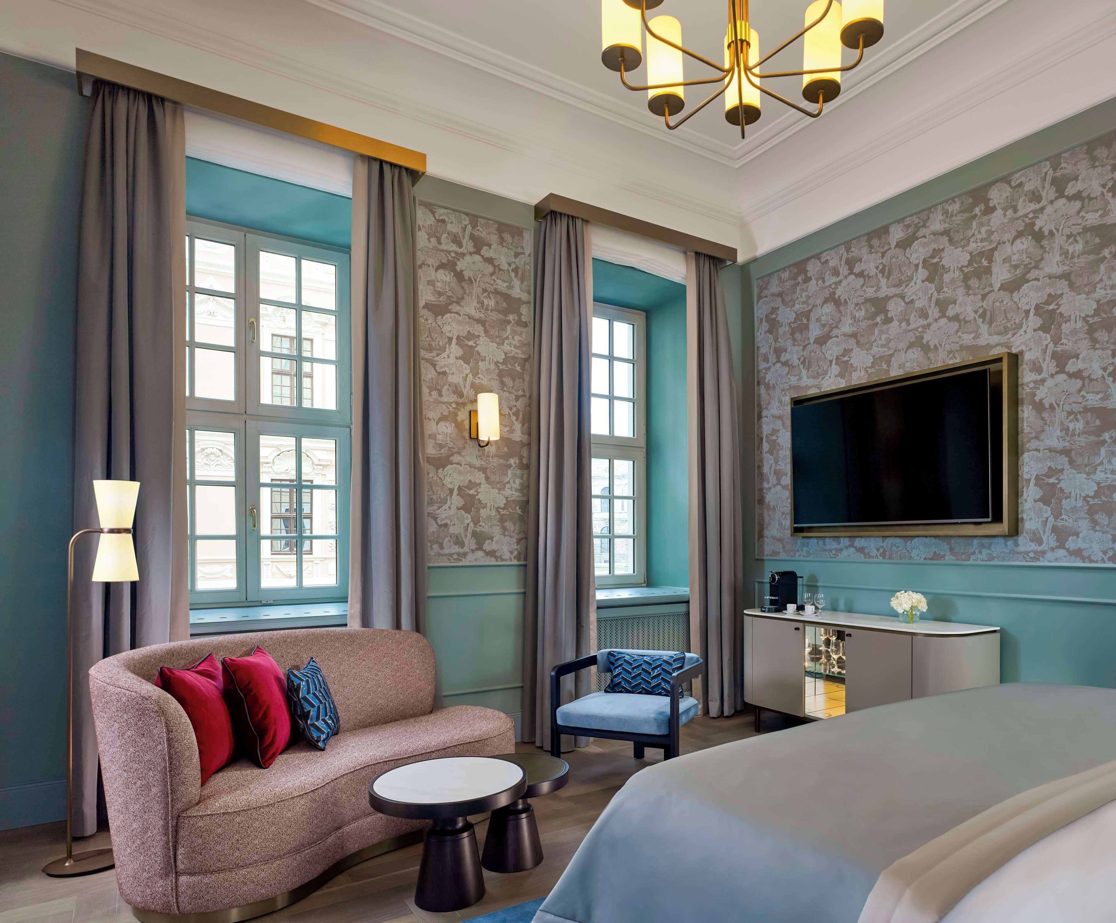 Ein Hotelzimmer mit einem großen Bett, einer Couch, einem kleinen runden Tisch, einem Sessel, einem Schreibtisch mit Flachbildfernseher und großen Fenstern mit Vorhängen.