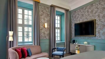 Ein Hotelzimmer mit einem großen Bett, einer Couch, einem kleinen runden Tisch, einem Sessel, einem Schreibtisch mit Flachbildfernseher und großen Fenstern mit Vorhängen.