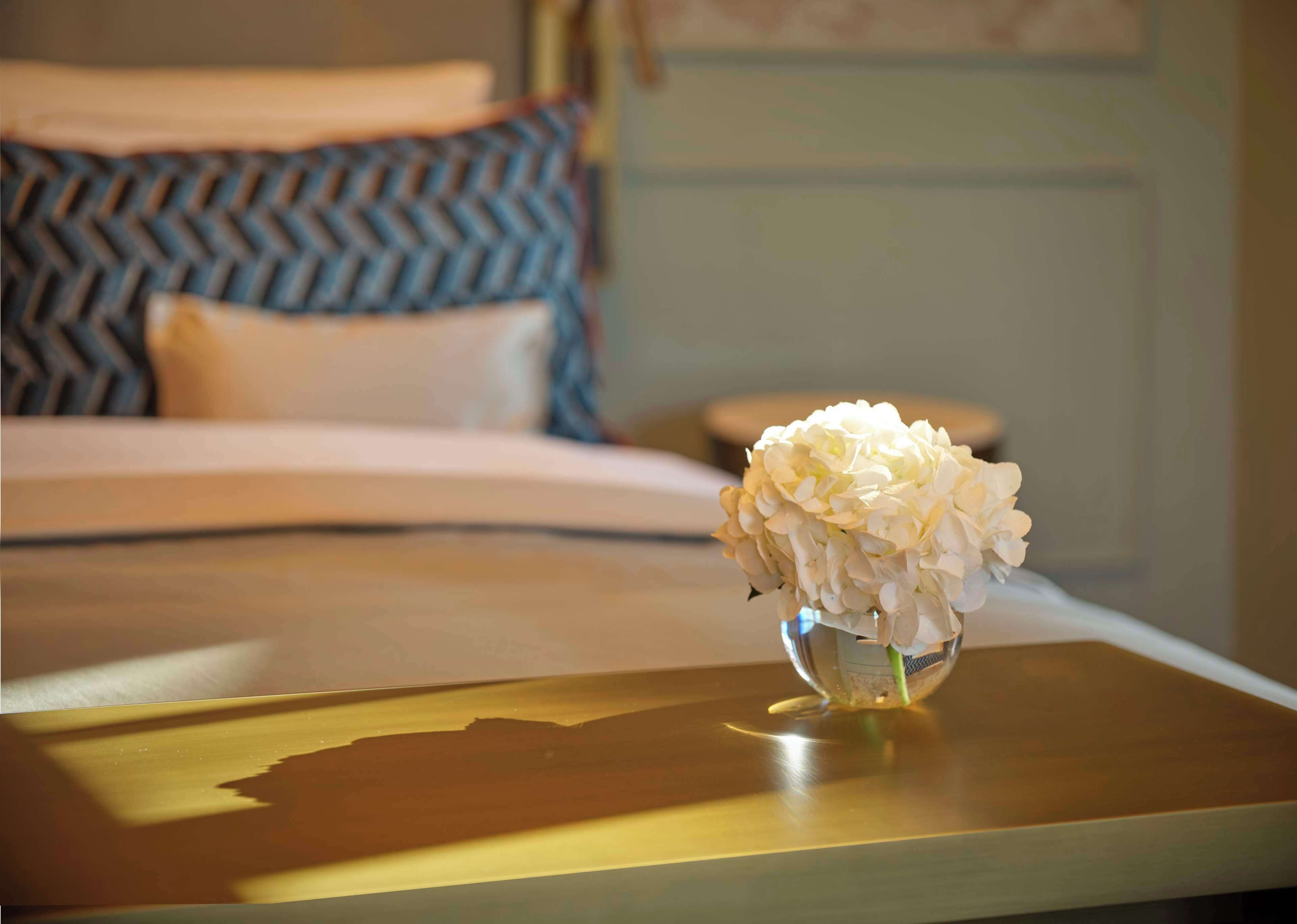 Ein Hotelzimmer mit einem Bett, einem Nachttisch mit einer Vase weißer Blumen und einem gemusterten Kissen.