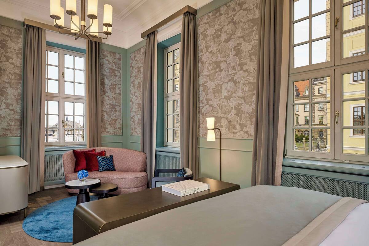 Ein Hotelzimmer mit einem Bett, einem Sofa, einem kleinen Tisch mit einem Buch, einer Lampe und Blick auf Gebäude durch große Fenster.