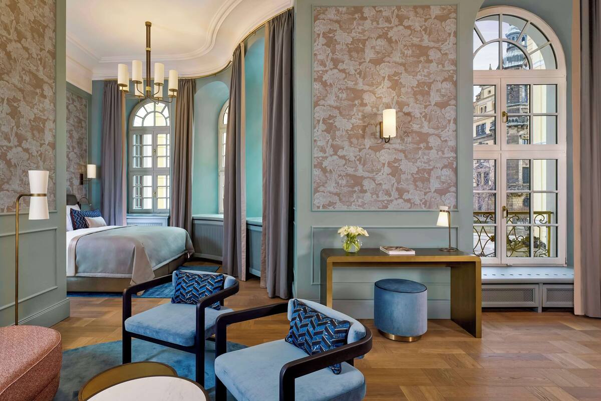 Ein Hotelzimmer mit einem großen Bett, zwei Sesseln, einem kleinen Tisch und einem Fenster mit Blick auf Gebäude.