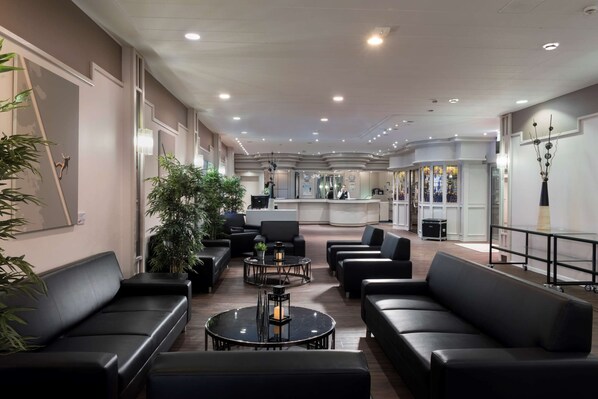 Relax in our spacious lobby, featuring a central seating area with comfortable sofas and round tables, a reception desk, and large windows offering views of the outside.
