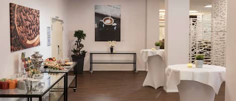 Un moderno vestíbulo de hotel con una mesa expositora de vidrio, un cuadro con temática de café, una obra de arte de taza de café, una planta en maceta y un separador blanco.