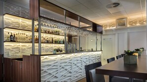 Un bar moderno con un original diseño de luces geométricas, una variedad de botellas de licor y una larga mesa de comedor con sillas.