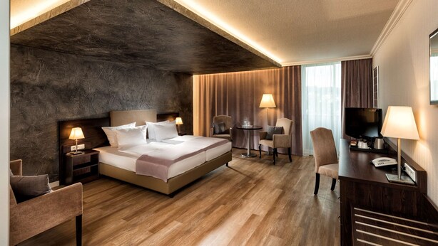 Una habitación de hotel moderna con una cama grande, mesitas de noche, un escritorio con silla, un televisor y una ventana con cortinas.