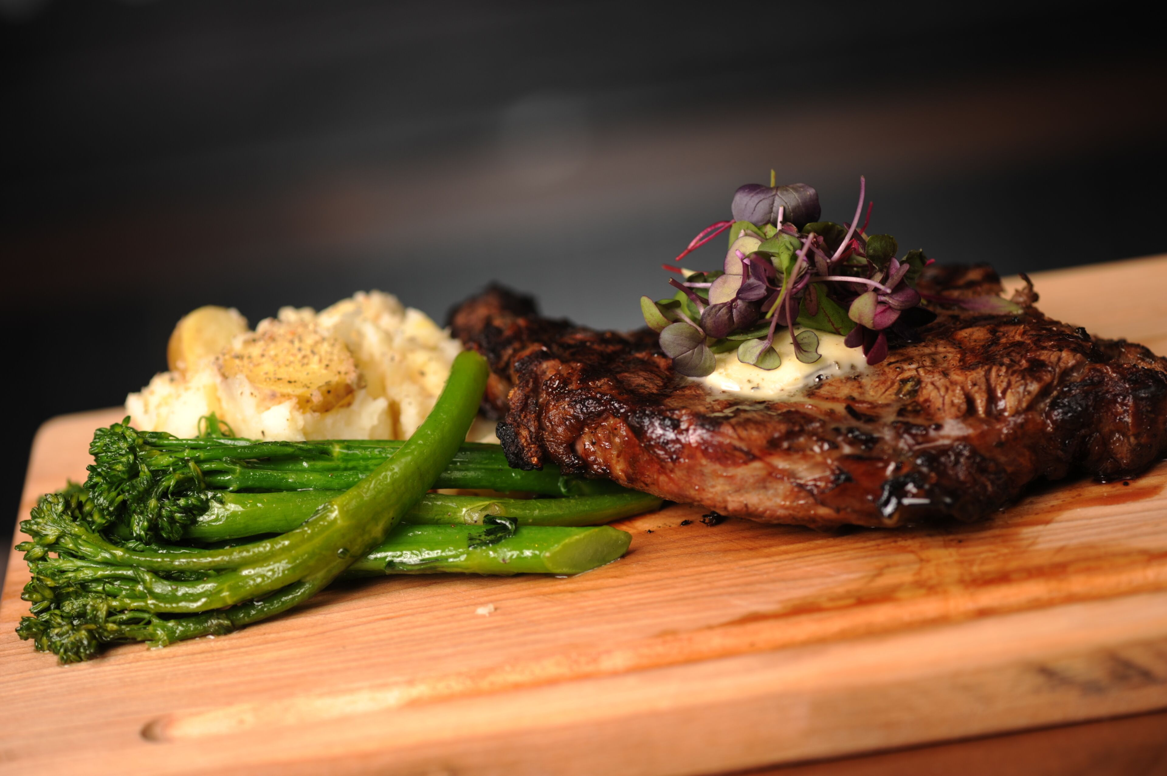 A steak with a garnish of greens and a side of mashed potatoes on a wooden board.