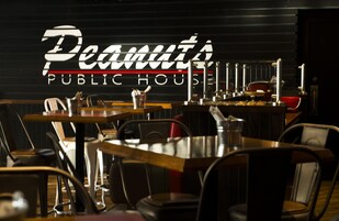 Interior de restaurante com mesas e cadeiras, um letreiro de neon com a inscrição "Peanut's Public House" e uma área de bar ao fundo.