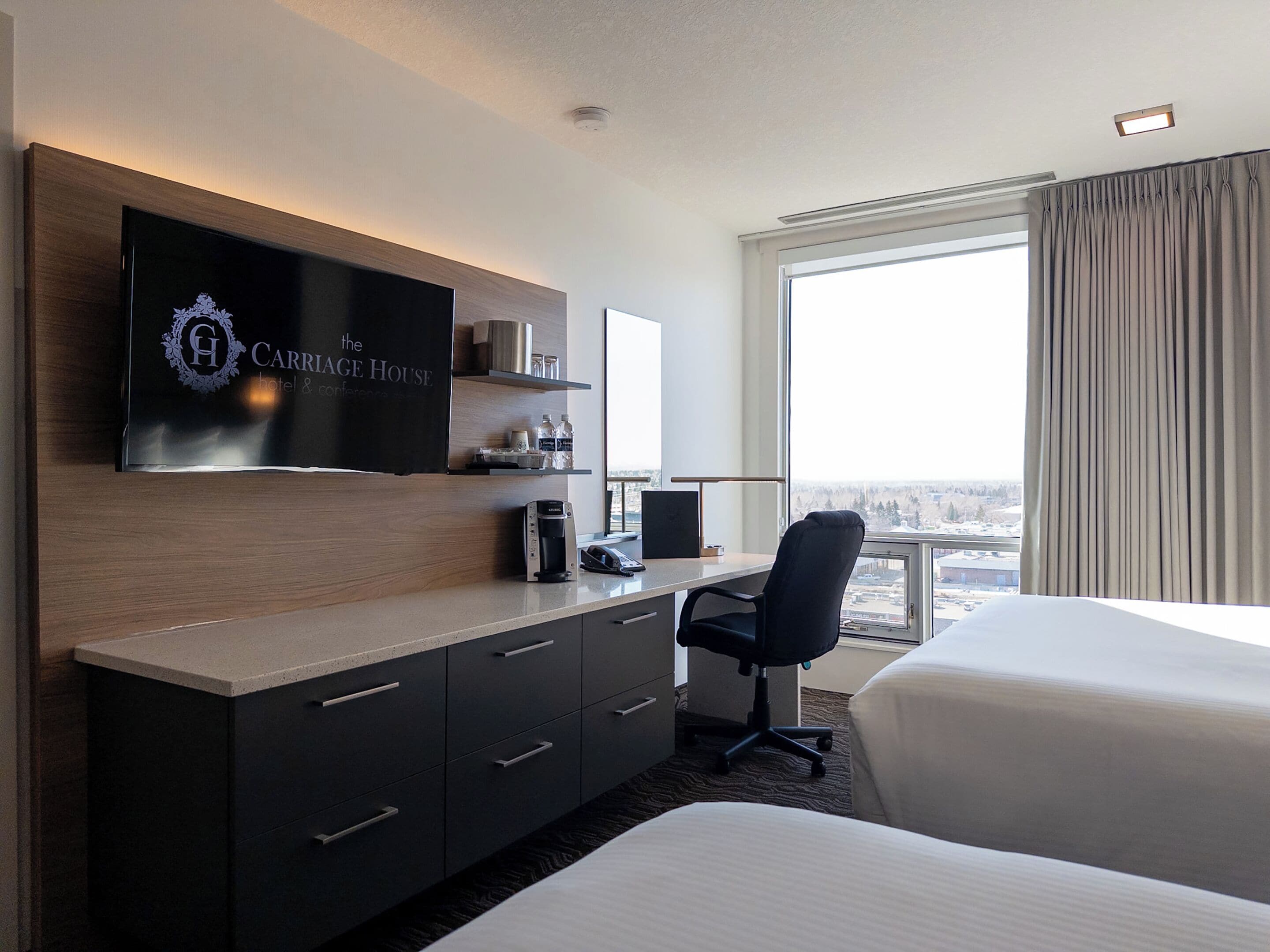 A hotel room with a flat-screen TV mounted on the wall, a desk with a chair, a bed, and a large window with curtains.