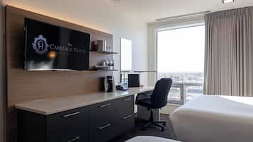 A hotel room with a flat-screen TV mounted on the wall, a desk with a chair, a bed, and a large window with curtains.