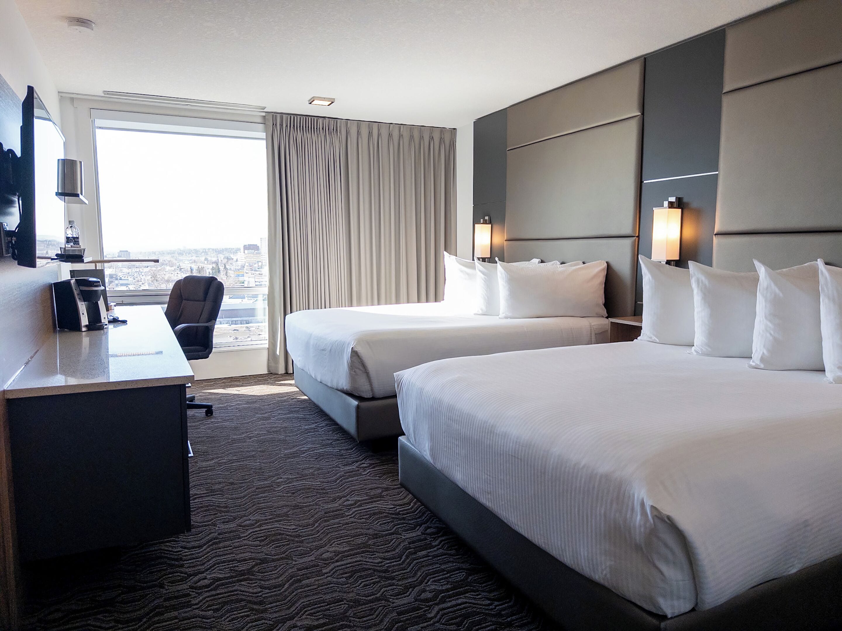 A hotel room with two beds, a desk, and a chair. There is a window with curtains, a view of the city, and a flat-screen TV on the wall.