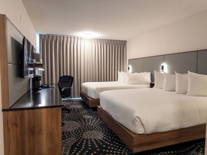 A hotel room with two beds, a desk with a flat-screen TV, a chair, and a large window with curtains.