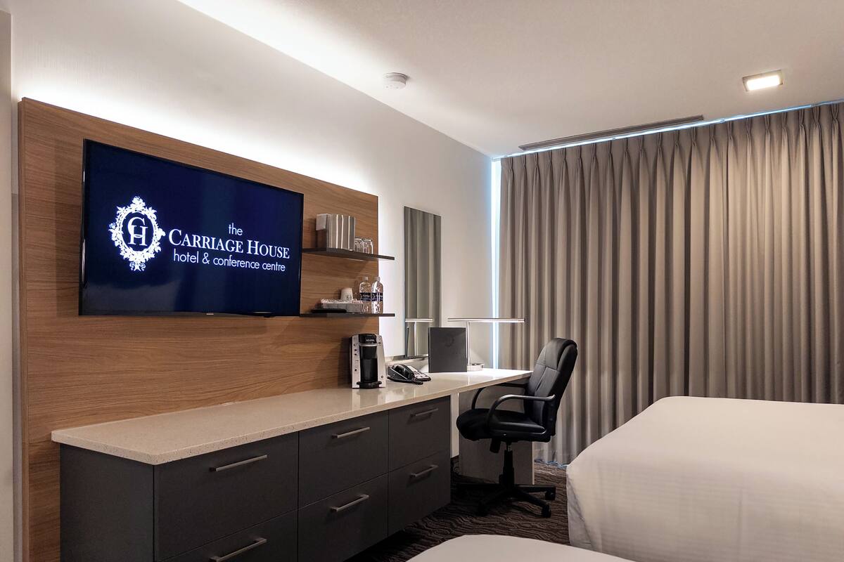 A hotel room with a bed, a desk with a computer, a chair, a TV mounted on the wall, and a minibar.