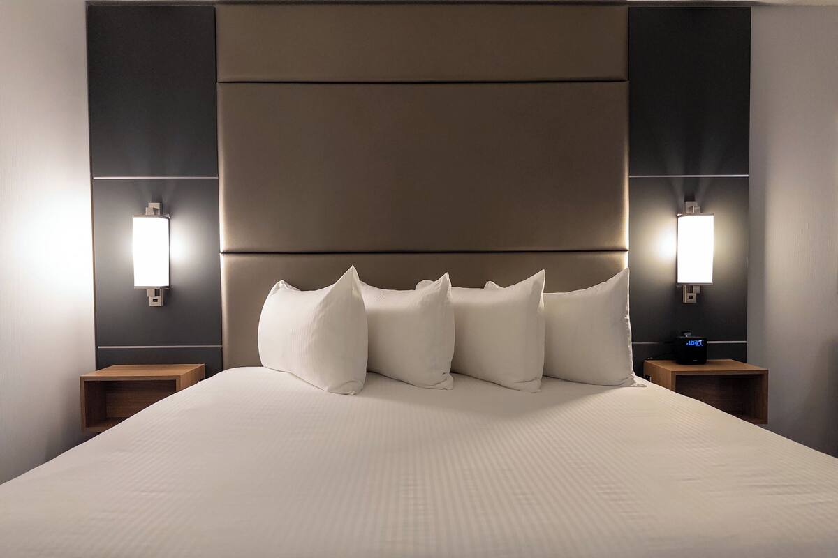A modern hotel room with a large bed, multiple pillows, and two bedside tables with lamps.