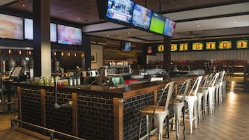 A bar area with multiple screens, a counter with taps and glasses, and rows of bar stools.