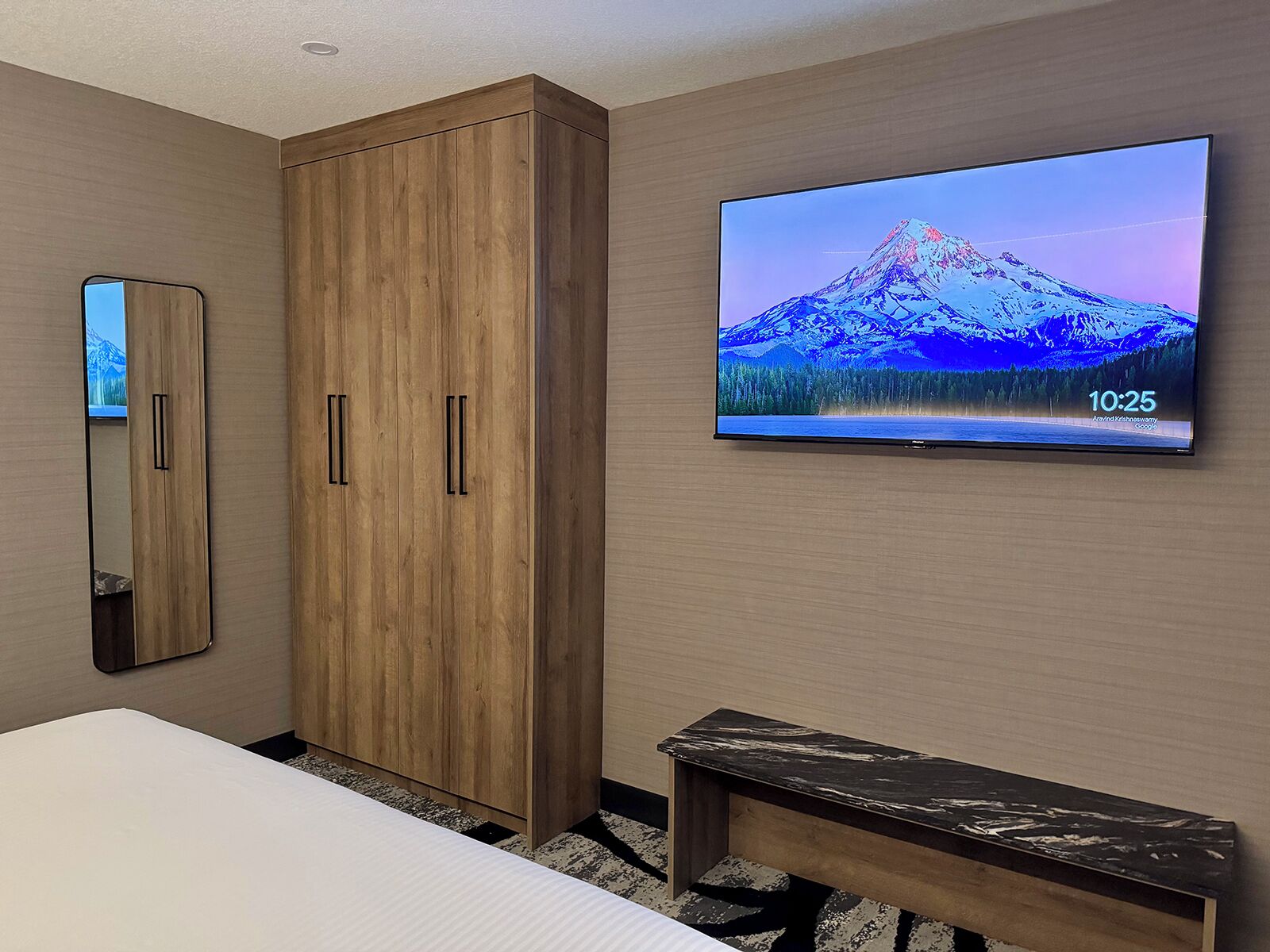 Habitación de hotel con un armario de madera, un televisor de pantalla plana grande mostrando un paisaje montañoso, una mesita de noche y un espejo.