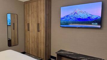 Habitación de hotel con un armario de madera, un televisor de pantalla plana grande mostrando un paisaje montañoso, una mesita de noche y un espejo.