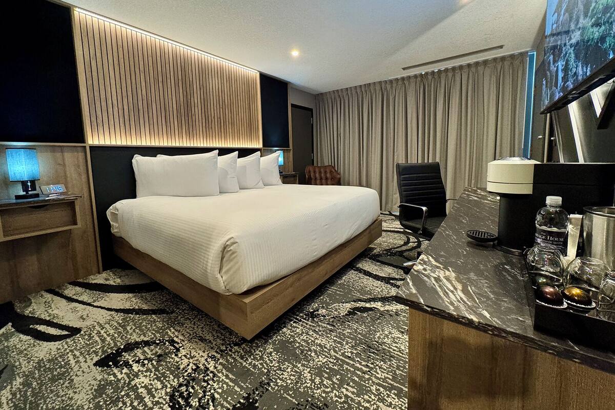 A modern hotel room with a large bed, a desk with a coffee maker, a chair, and a large window with curtains.