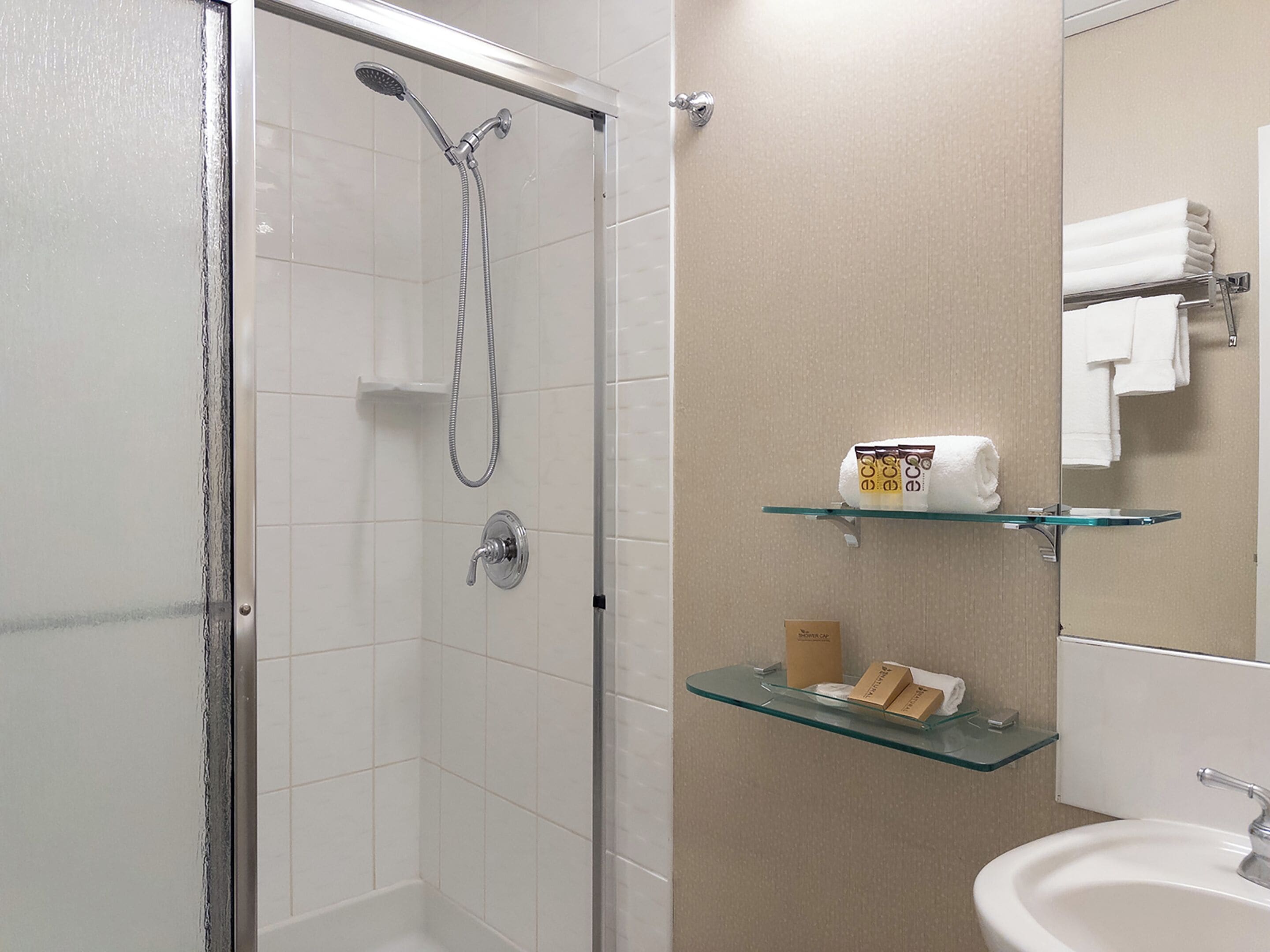 Een badkamer met een douchecabine van glas, een glazen plank met toiletartikelen, en een wastafel met een spiegel erboven.