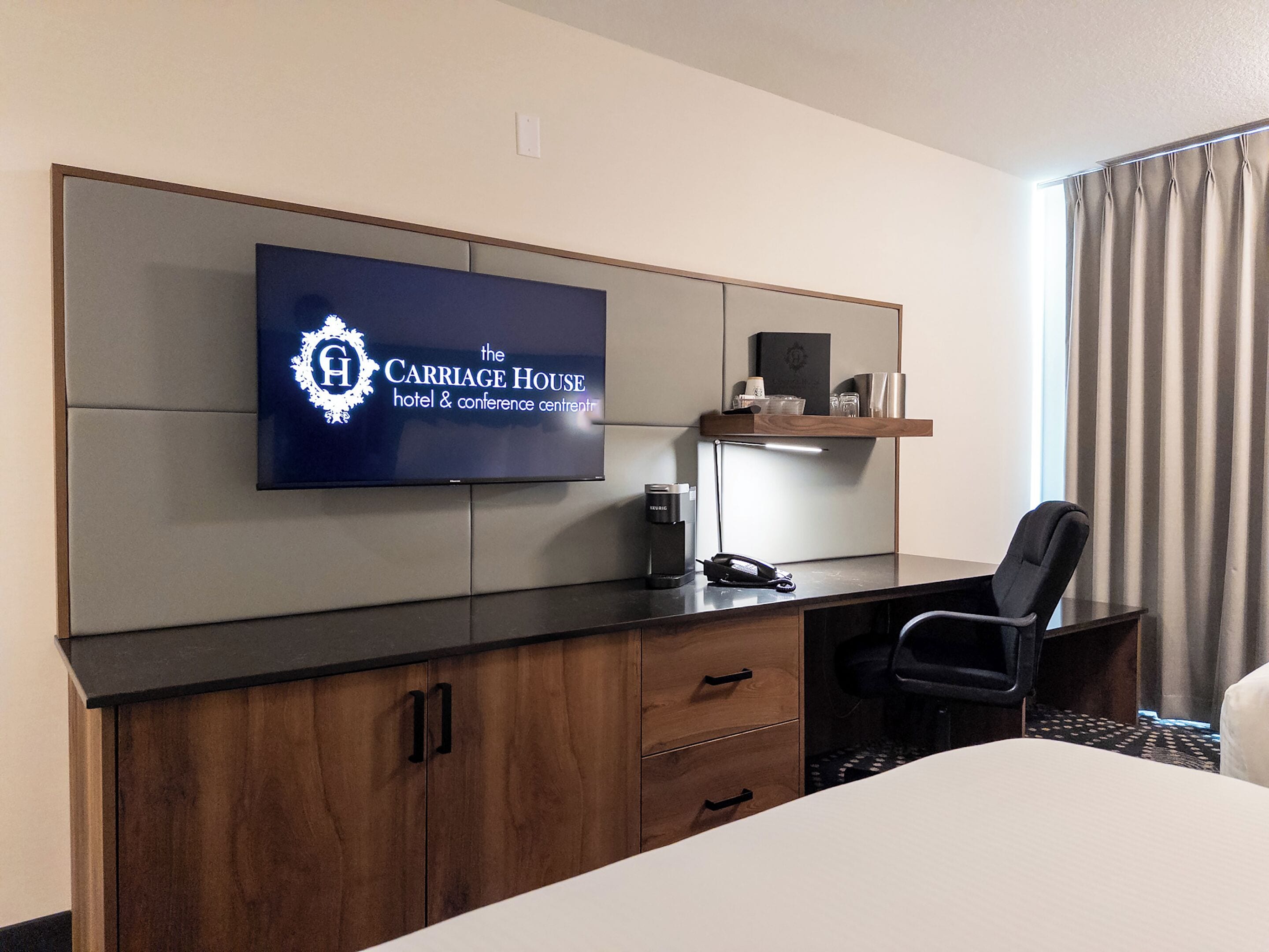 A hotel room with a large flat-screen TV mounted on the wall, a wooden desk with a chair, a minibar, and a bed with a white comforter.
