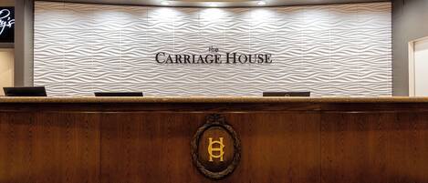 Un comptoir d’accueil avec un devant en bois sculpté, un luminaire circulaire au plafond et un mur sur lequel est inscrit le texte « The Carriage House ».