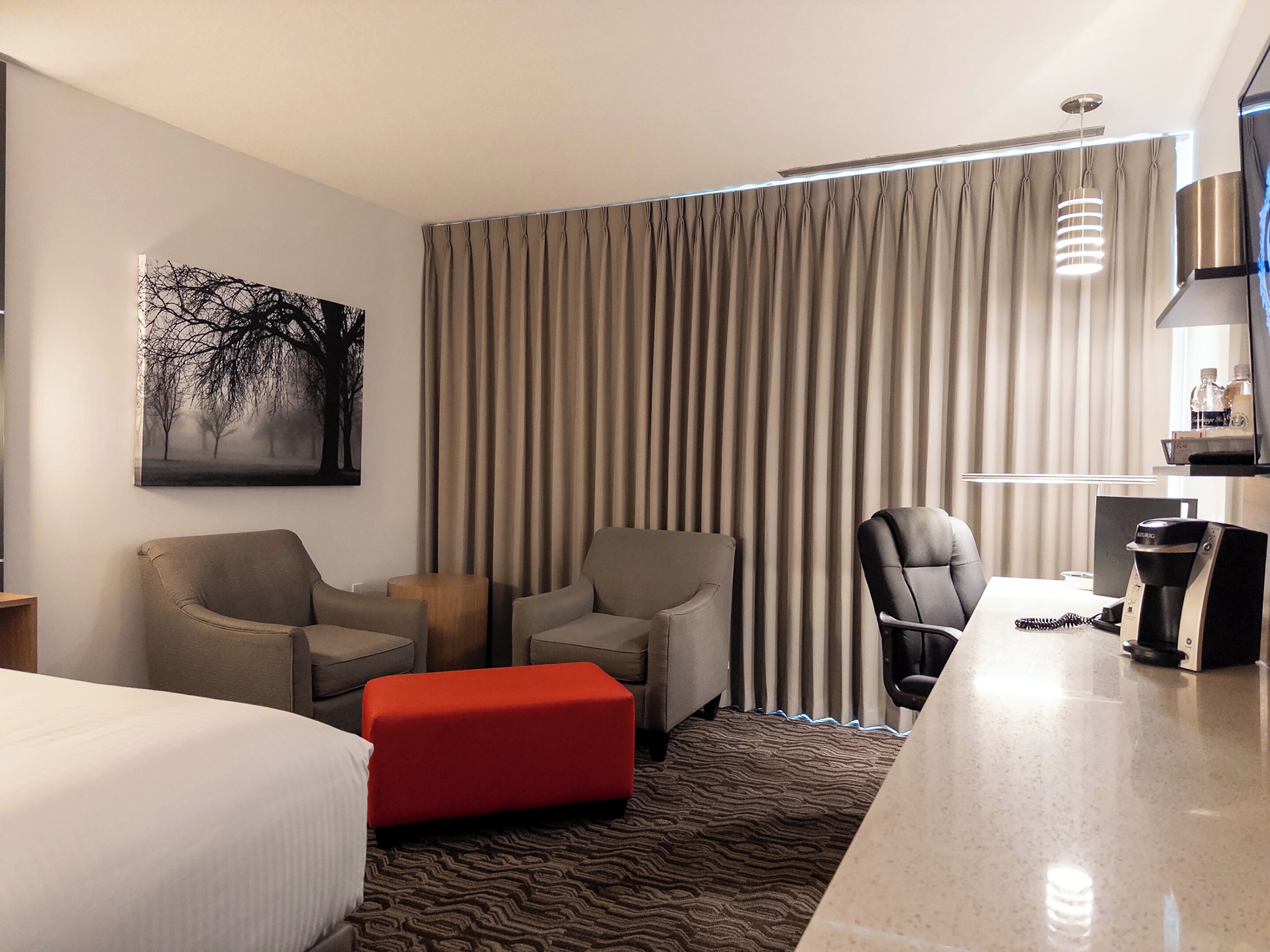 A hotel room with a bed, two armchairs, a red ottoman, a desk with a coffee machine, and a large window with curtains.