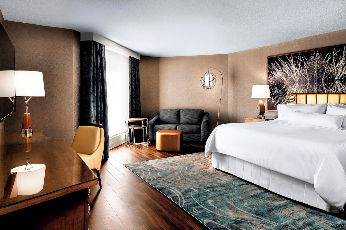 A hotel room with a large bed, a desk with a lamp, a chair, a small table, and a large window with curtains.