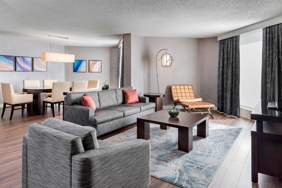 A modern living room with a grey sofa, a coffee table, and a TV. There are chairs, a rug, and artwork on the walls.