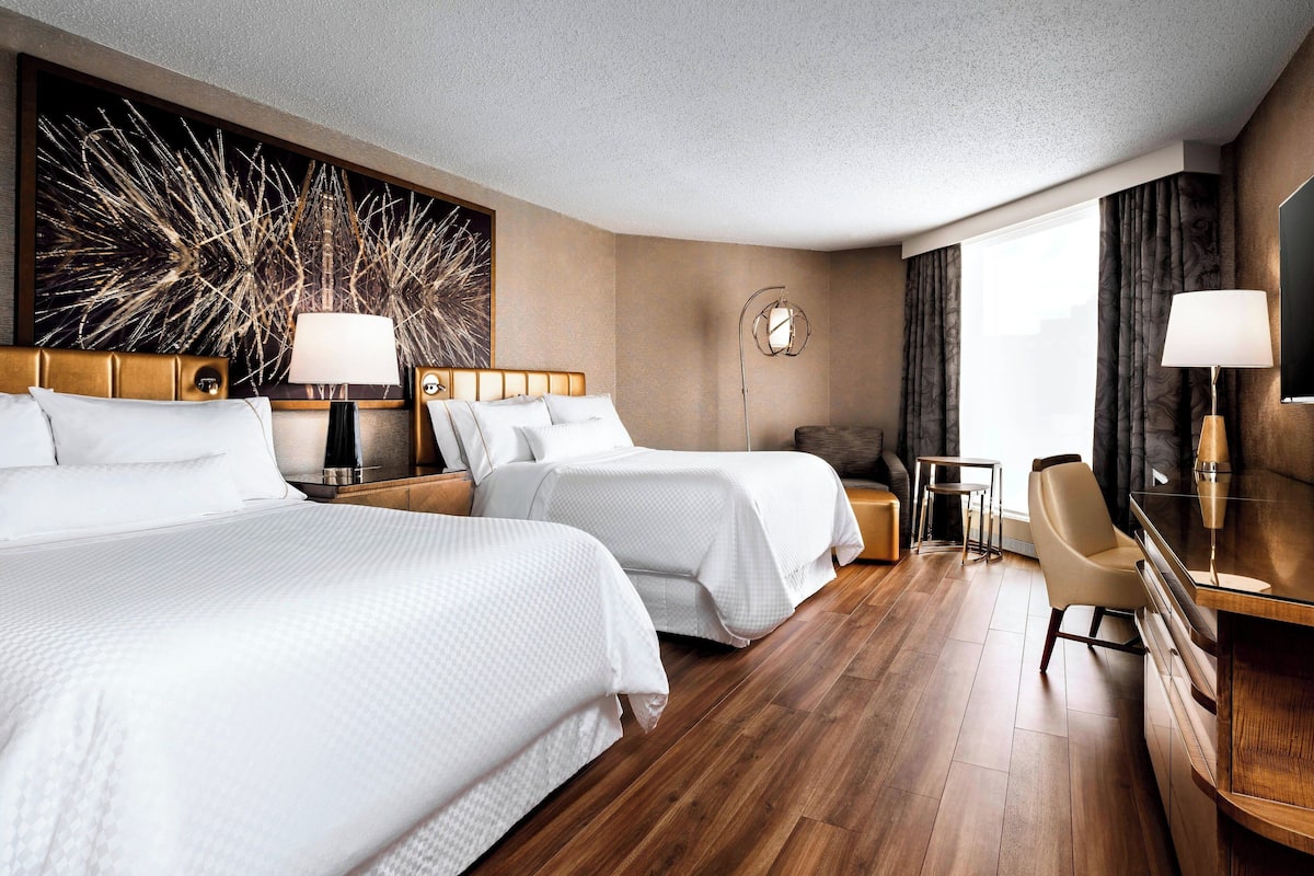 A hotel room with two beds, a desk, a chair, a large abstract painting, and a view of the outdoors.