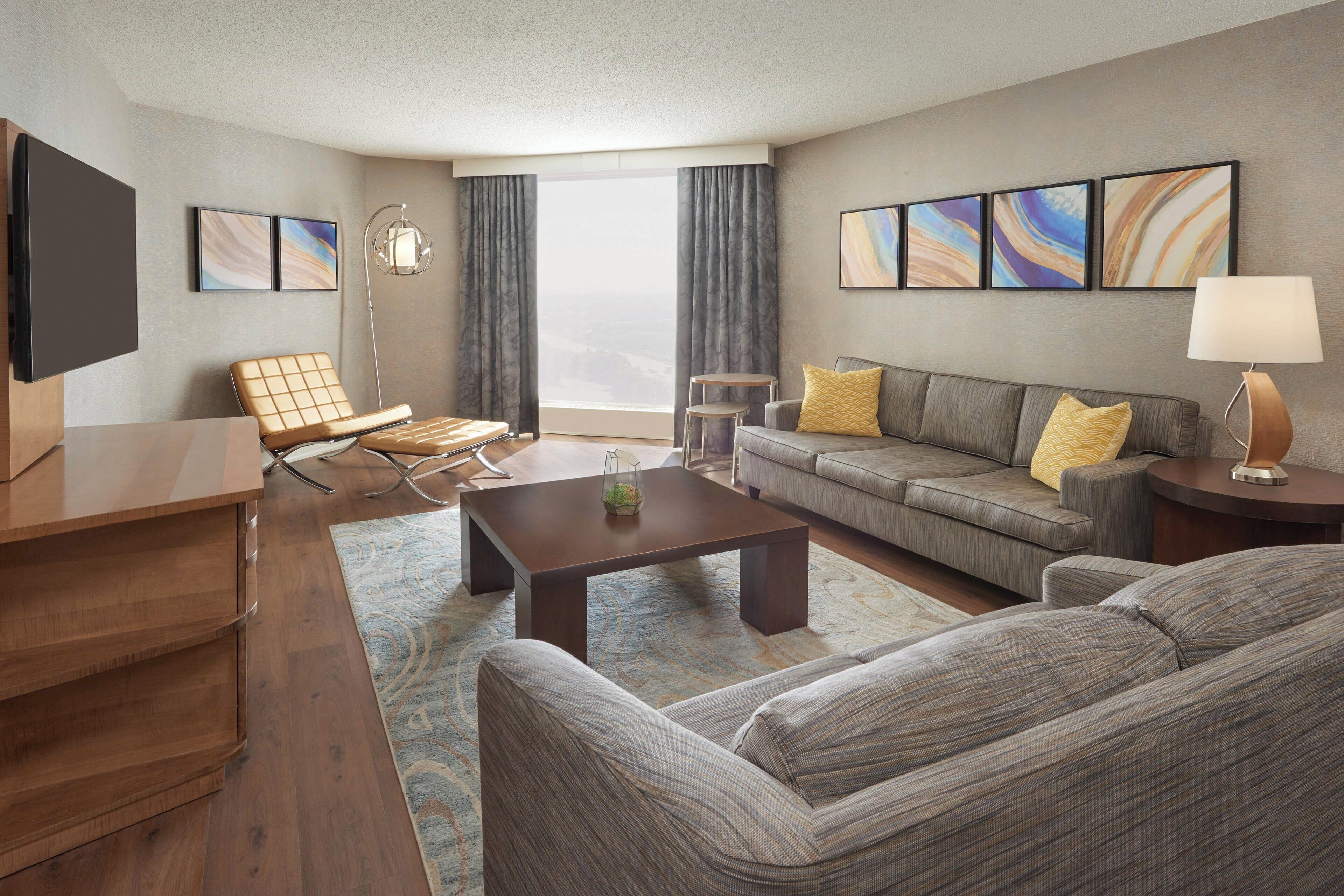 A modern hotel room with a sofa, armchair, coffee table, flat-screen TV, and artwork on the walls.