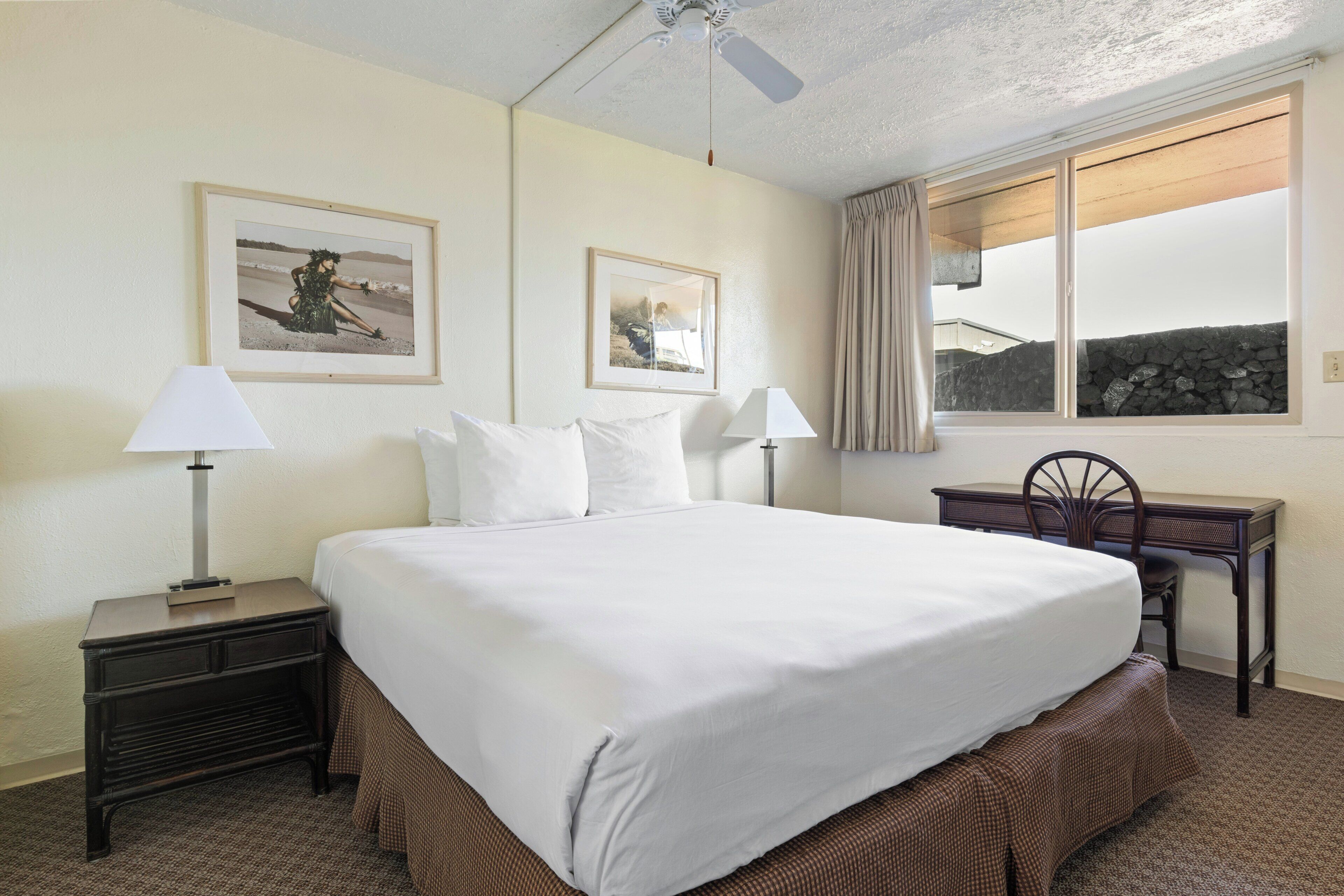 A hotel room with a bed, two bedside lamps, a desk with a chair, and two framed pictures on the wall.