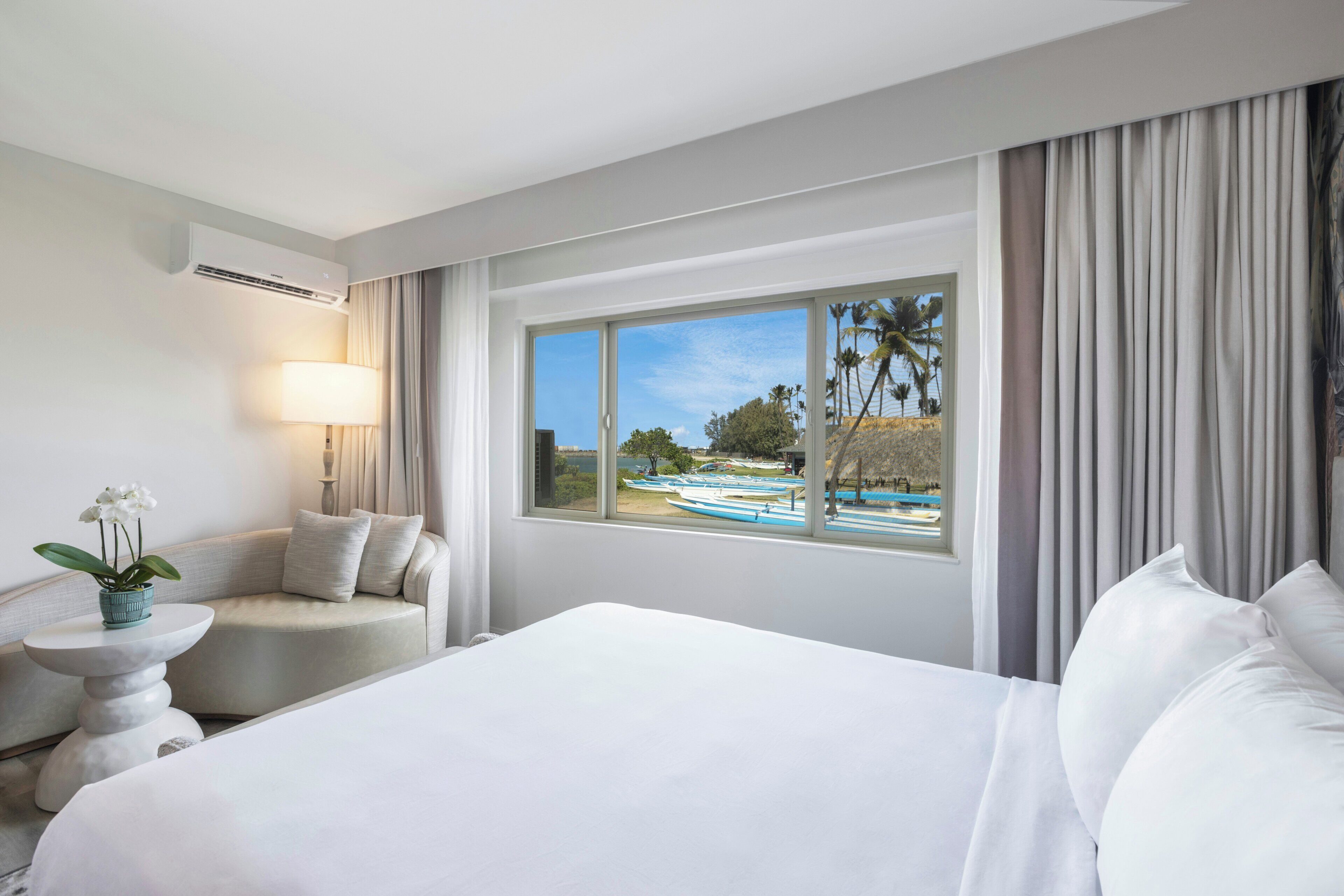 A hotel room with a large bed, a seating area with a lamp, and a window with a view of boats and palm trees.