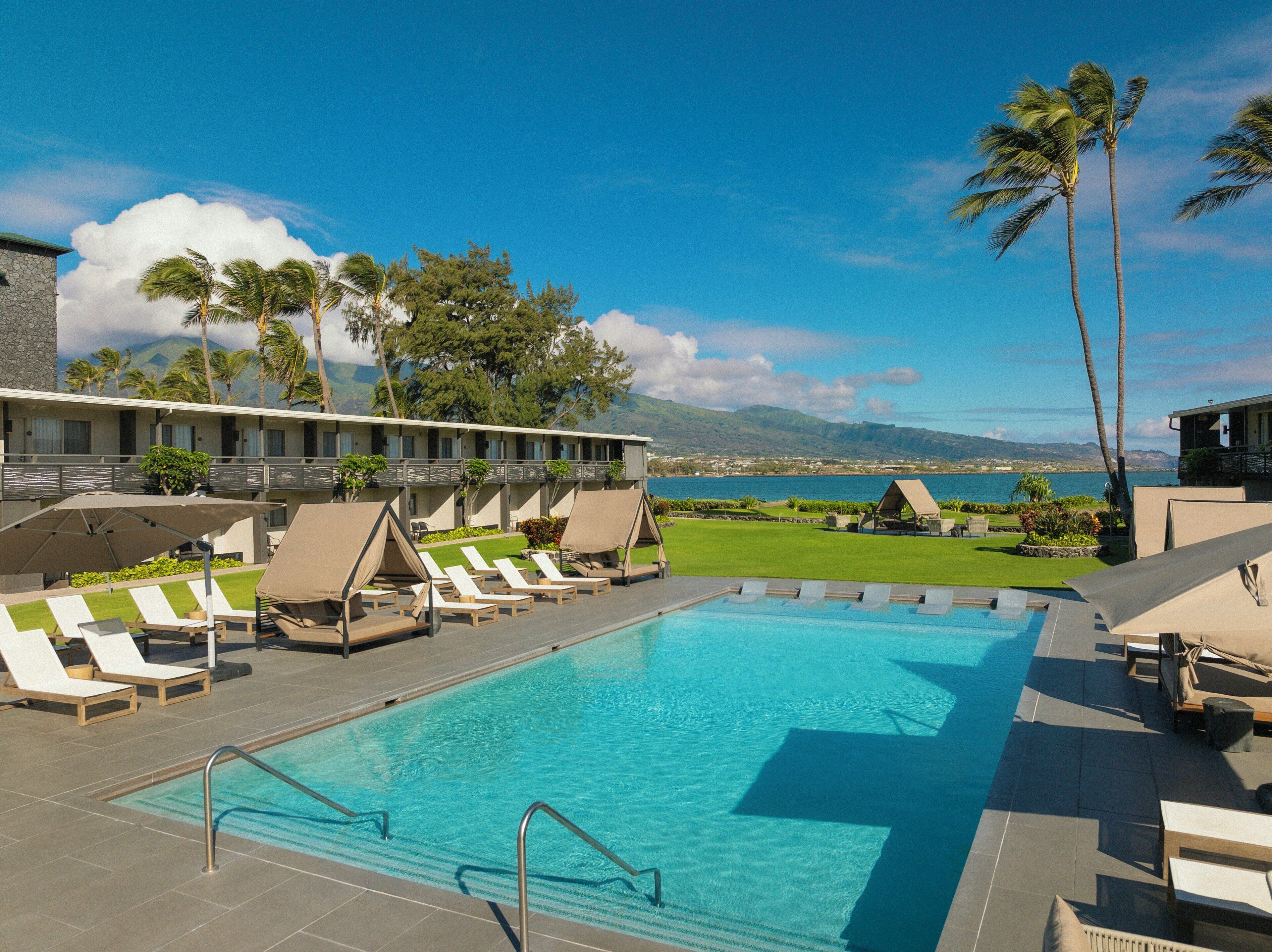 A pool area with lounge chairs, a clear blue pool, and a view of the ocean and mountains.