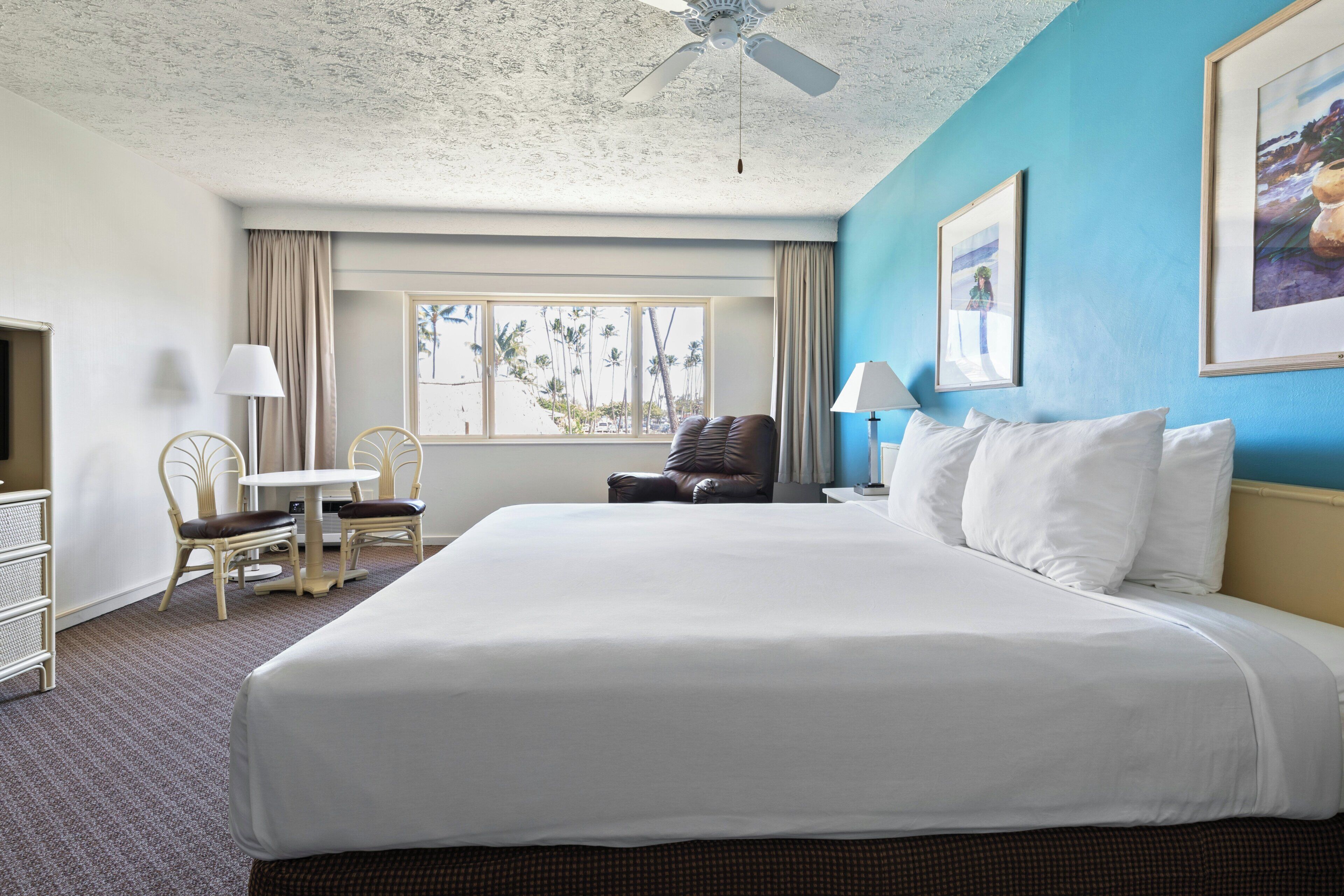 Habitación de hotel con una cama grande, un escritorio con silla, ventilador de techo y ventana con vista a palmeras.