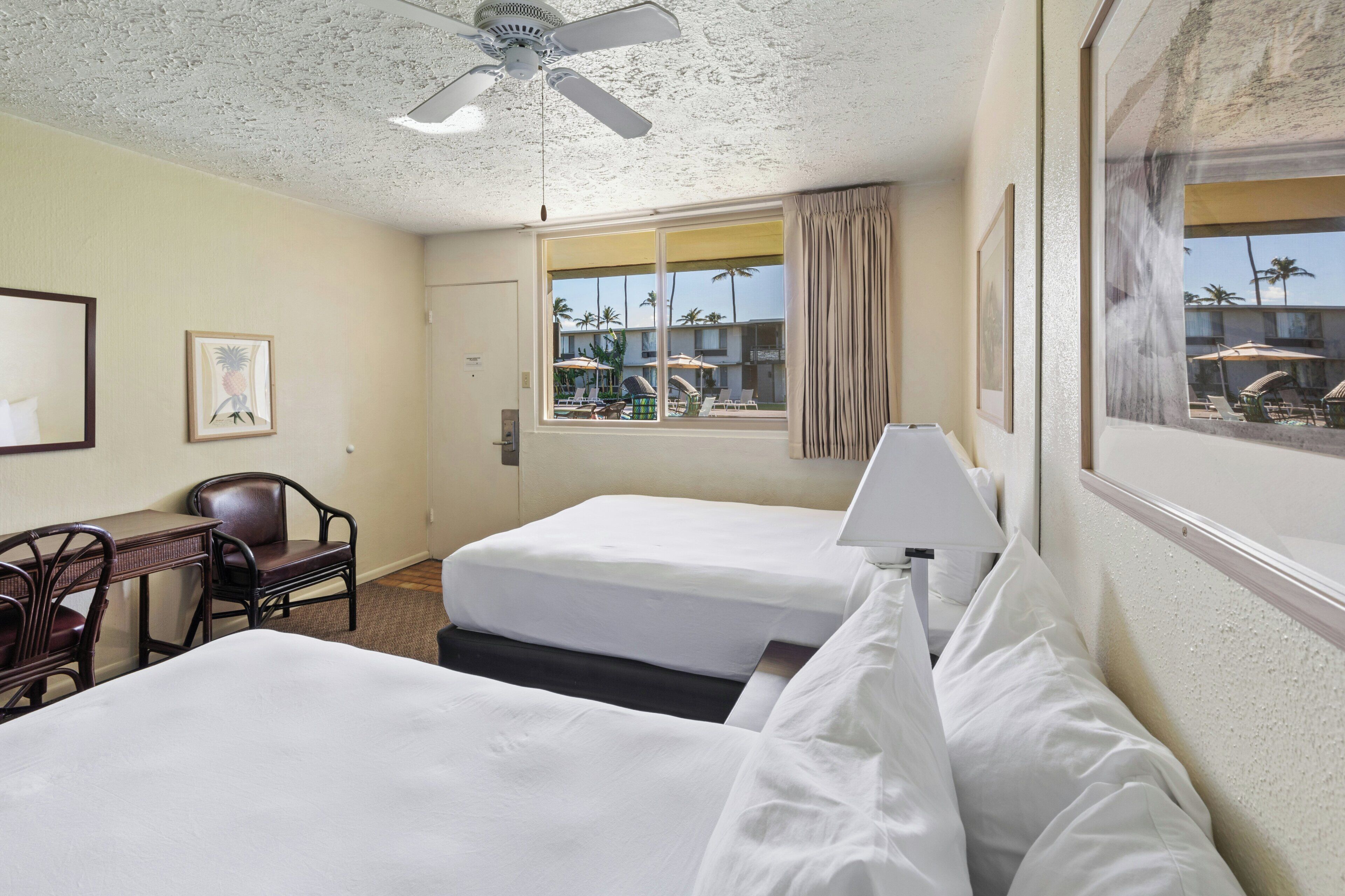 Une chambre d’hôtel avec deux lits, un bureau avec une chaise, un ventilateur de plafond et une fenêtre donnant sur des palmiers.