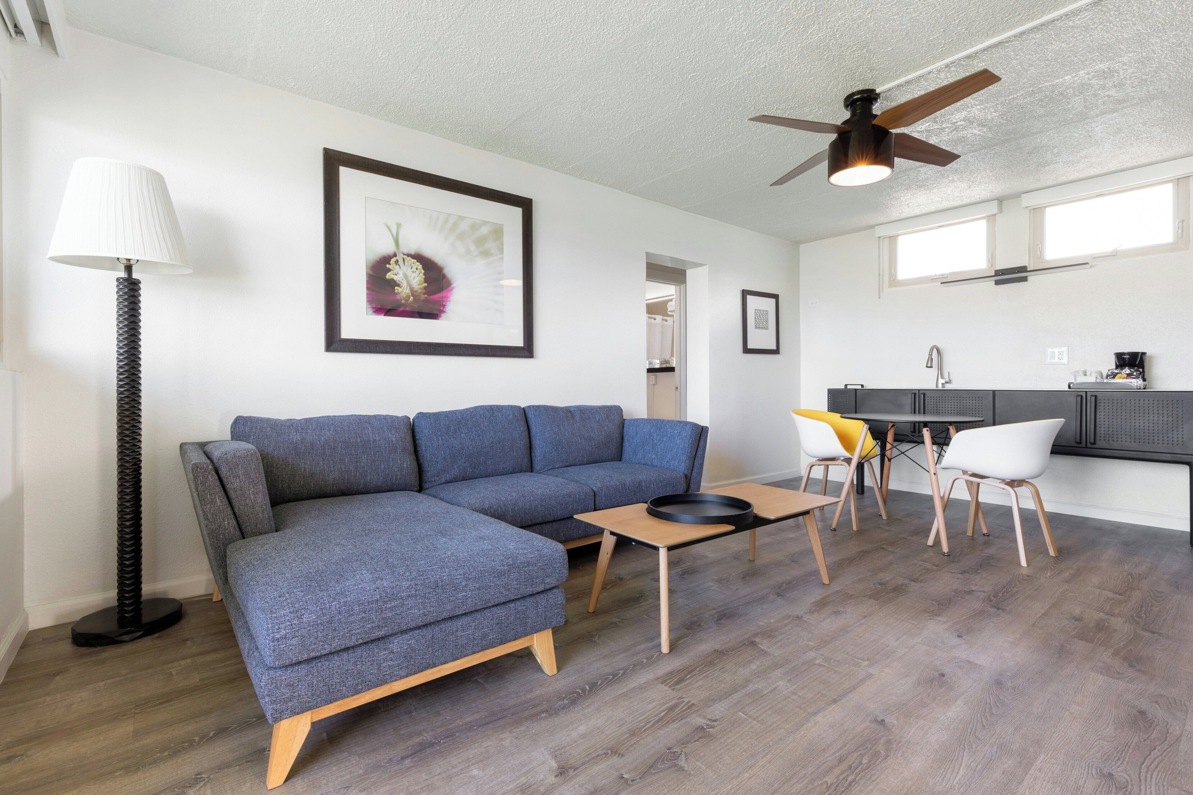 A modern living room with a sectional sofa, a coffee table, a floor lamp, a ceiling fan, and a kitchen area with a dining table and chairs.