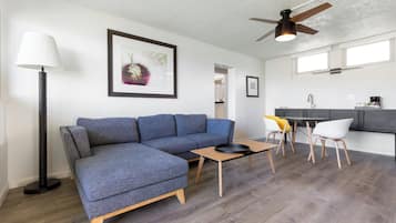 Un salon moderne comprenant un canapé d’angle, une table basse, un lampadaire, un ventilateur de plafond et un coin cuisine avec une table à manger et des chaises.