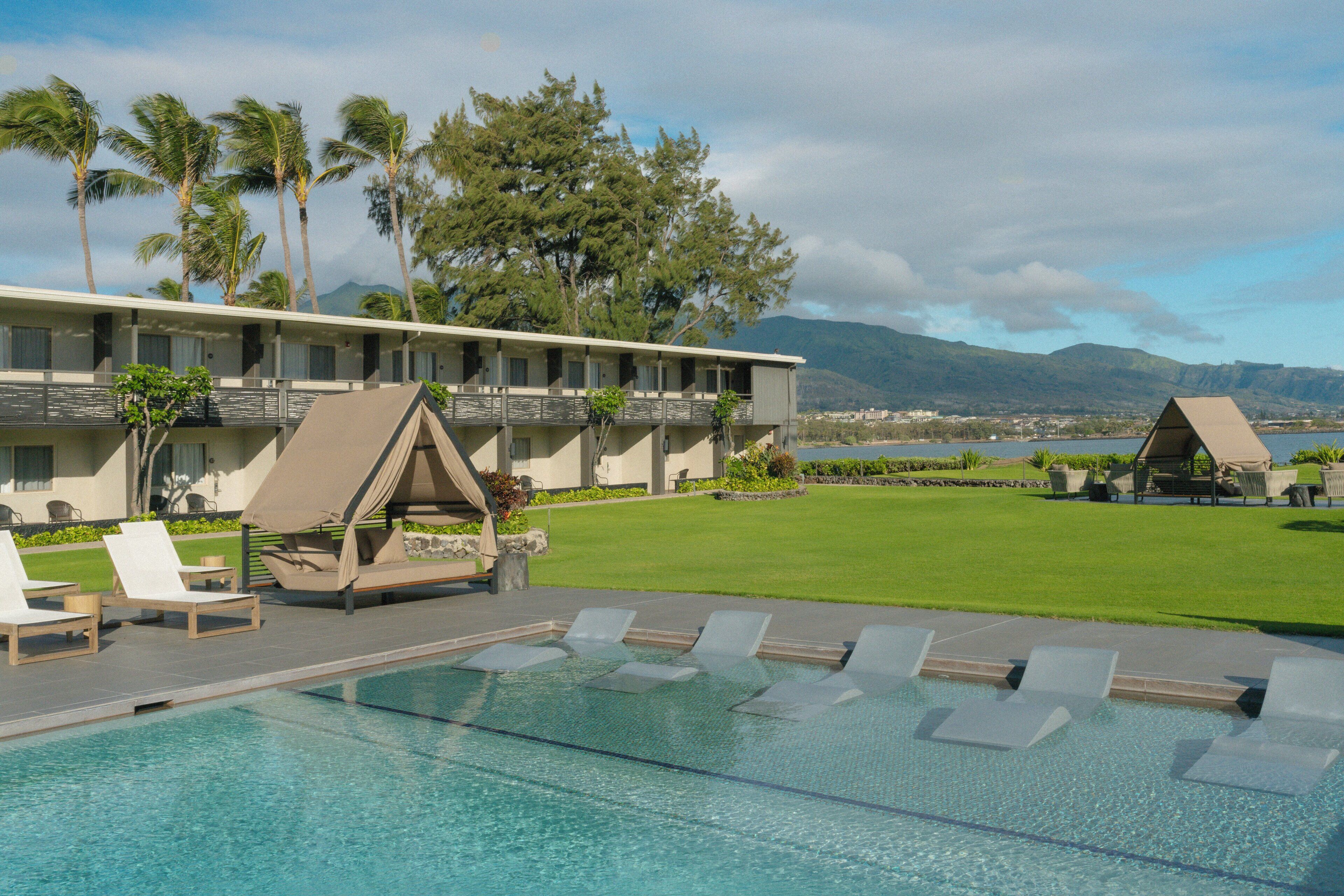 A hotel with a swimming pool, lounge chairs, and a view of mountains and a body of water.