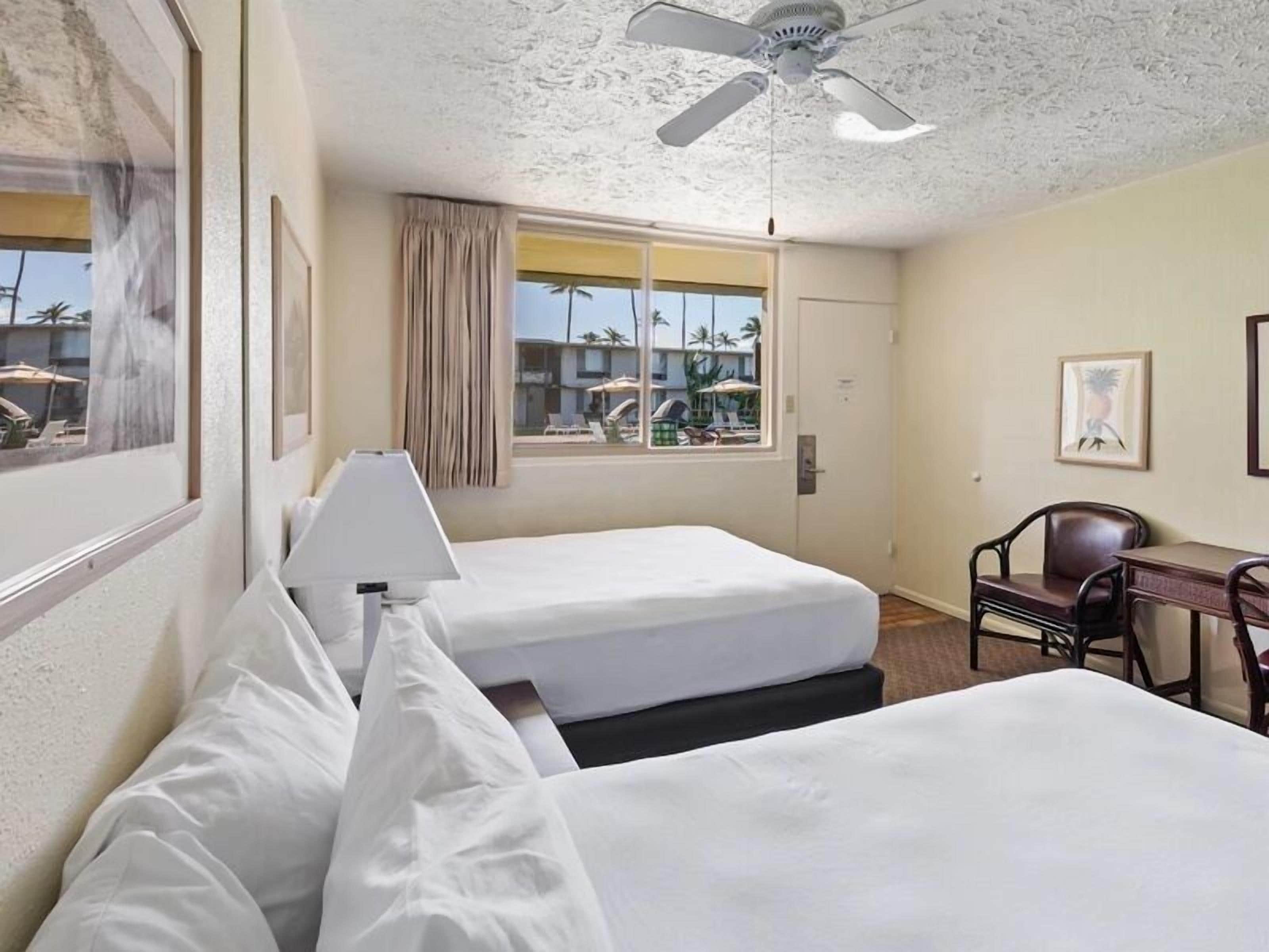 A hotel room with two beds, a ceiling fan, a window with a view of palm trees, and a desk with a chair.