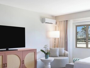 A hotel room with a television, a pink cabinet with a circular design, a round white table with a plant, a sofa with cushions, and a window with a view of palm trees. - Maui Seaside Hotel (Kahului)