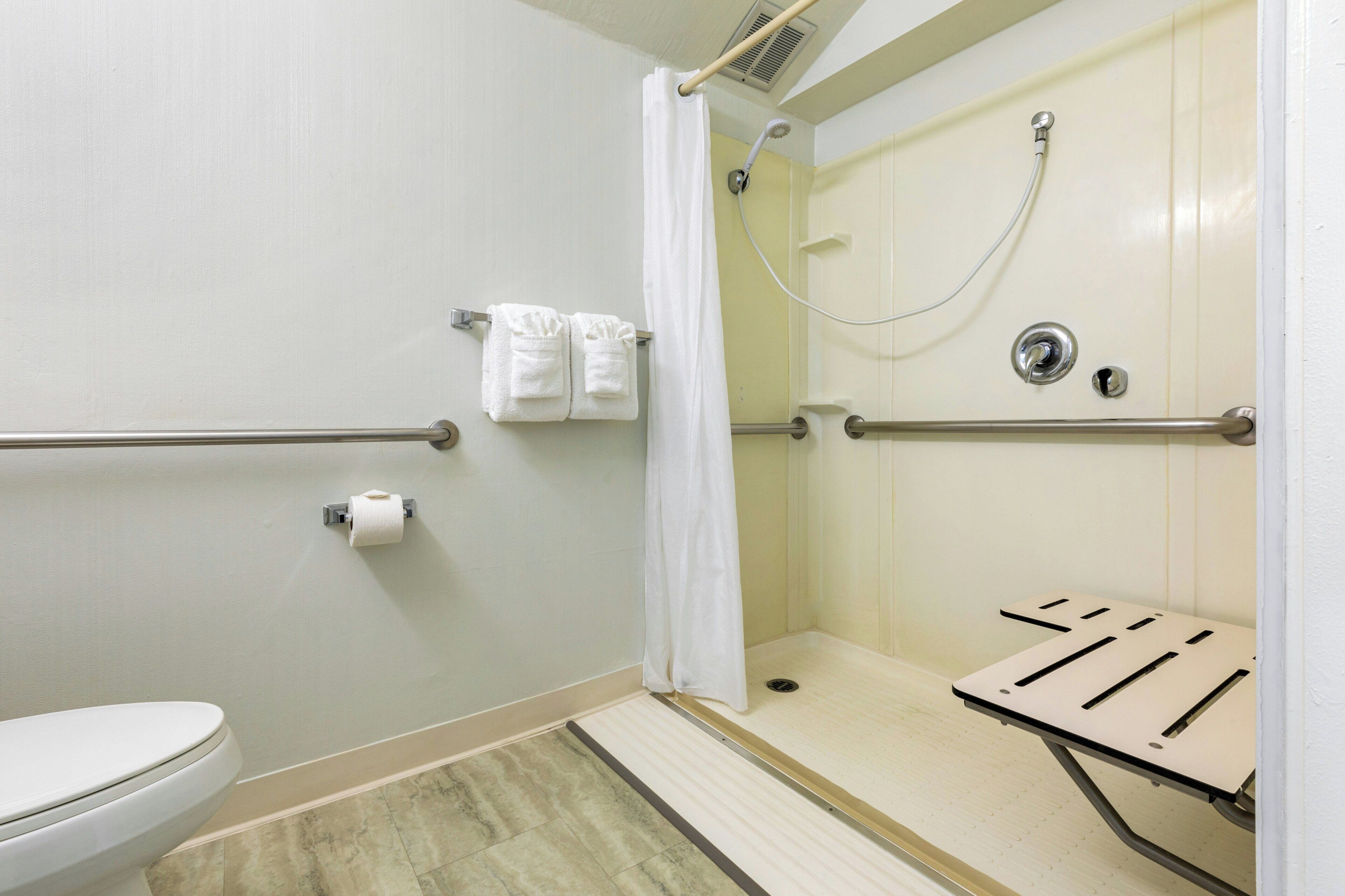 Ducha accesible en silla de ruedas con barra de apoyo, inodoro y rollo de papel higiénico.