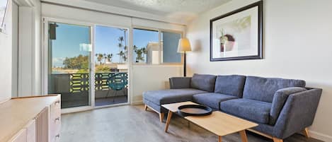 En moderne stue med en grå hjørnesofa, et træbord og en skydedør, der fører ud til en balkon med havemøbler.