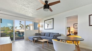 A modern living room with a grey sofa, a coffee table, and a dining table with chairs. There is a ceiling fan, a balcony with a view of palm trees, and a large window. - Maui Seaside Hotel (Kahului)