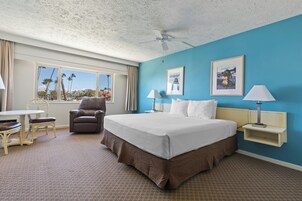 A hotel room with a large bed, a desk with a chair, a window with a view of palm trees, and two wall-mounted lamps.