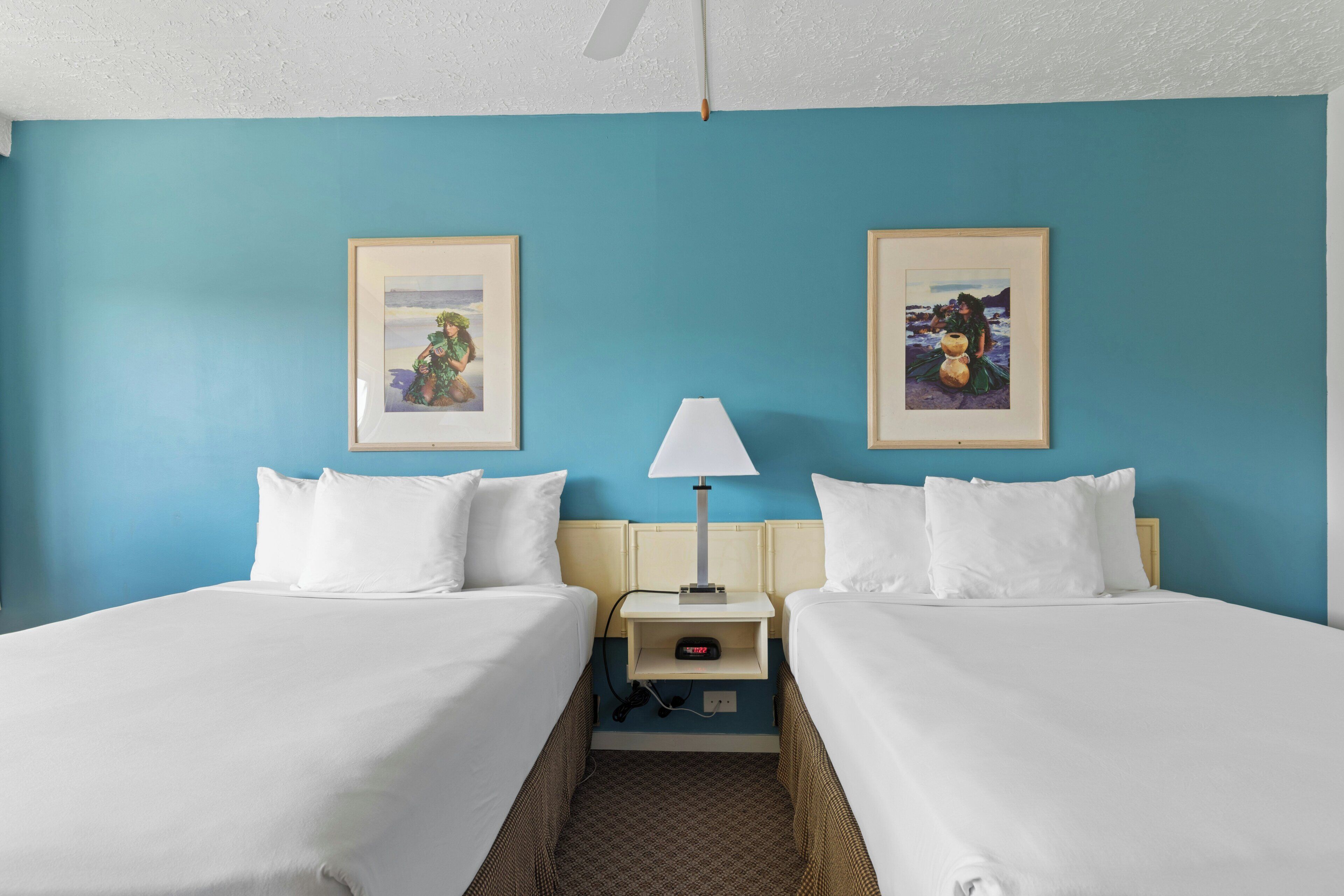 A hotel room with two beds, a nightstand with a lamp, and framed pictures on the walls.