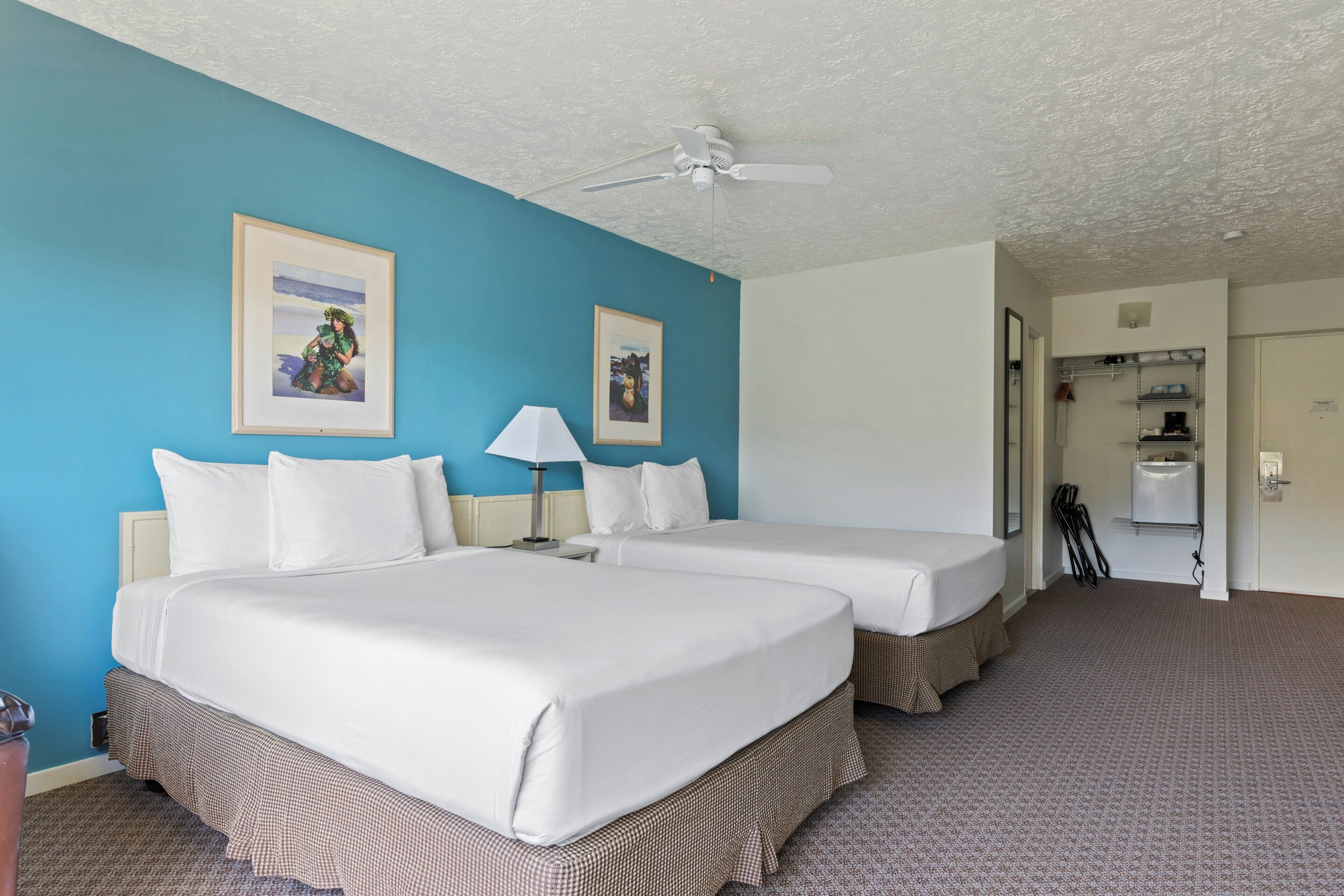 A hotel room with two beds, a ceiling fan, framed pictures on the wall, a lamp, and a bathroom visible through an open door.